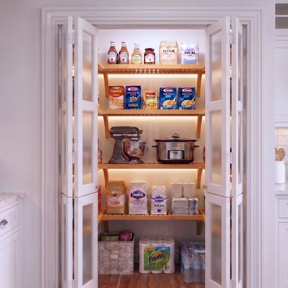 A small U shaped walk-in pantry with four levels of honey maple solid wood shelving filled with grocery items.