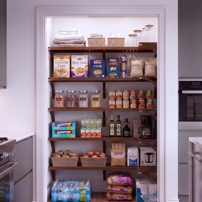 A small L shaped walk-in pantry with 5 levels of chestnut solid wood shelving filled with grocery items.