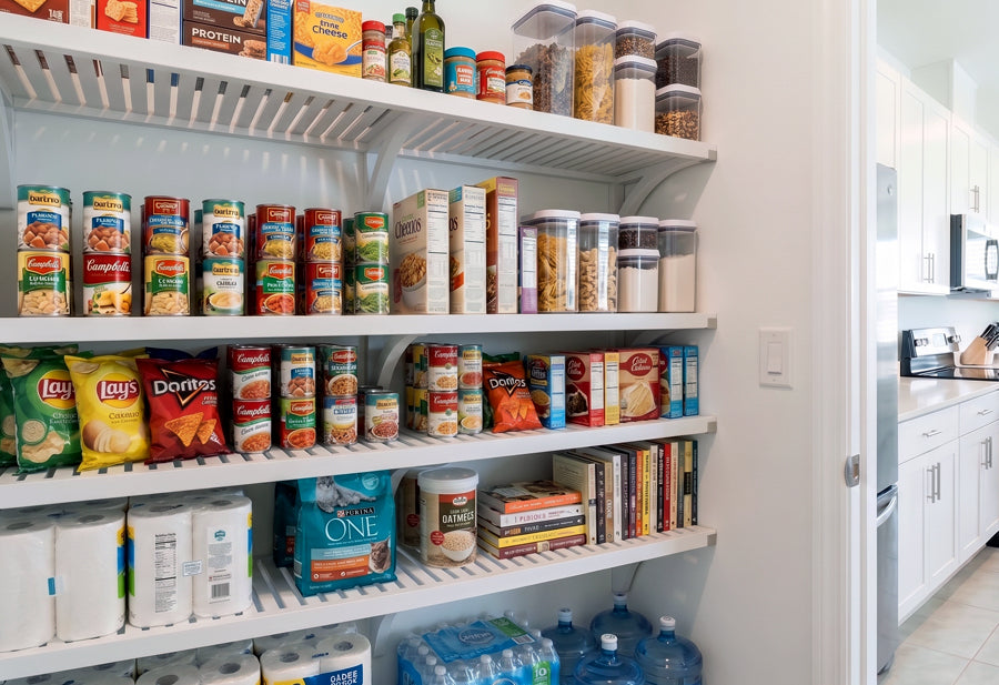 An after image of a  well-stocked white pantry using solid wood shelving filled with grocery essentials, with a white kitchen visible in the background.